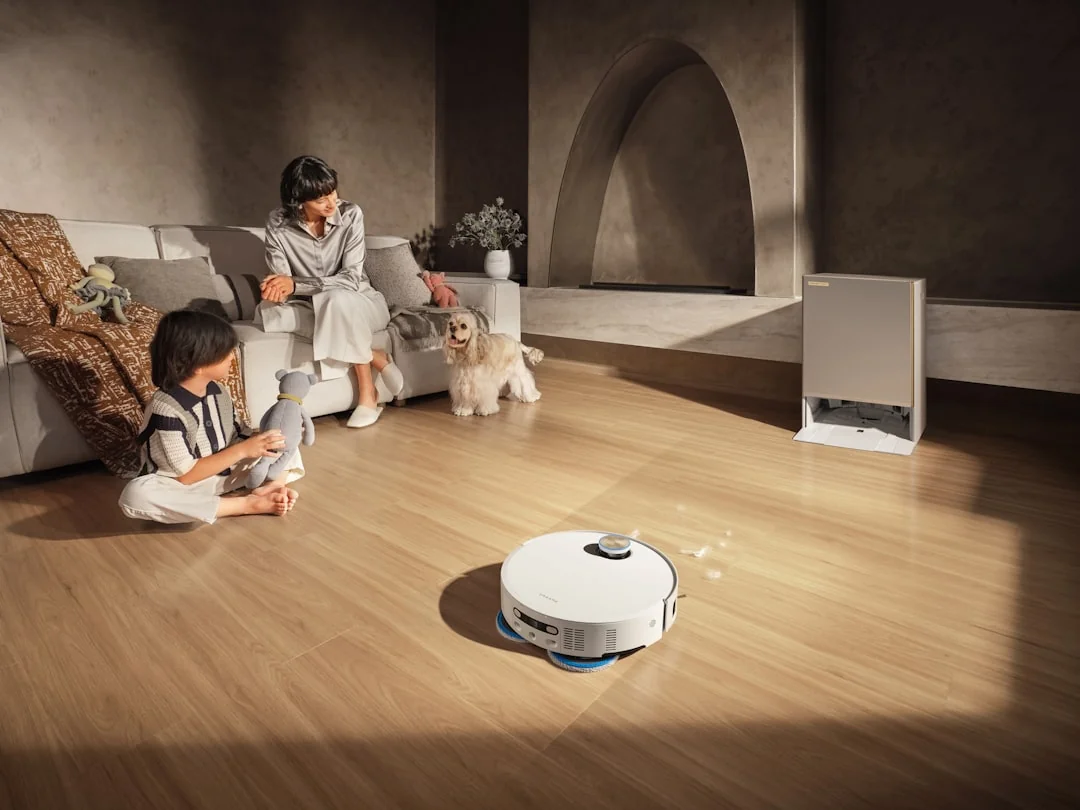 A woman vacuuming the floor while a dog looks on attentively. Indoor home setting.